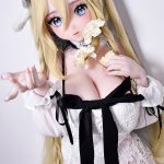 Cosplay doll with long blonde hair in a white lace dress, holding pale flowers and gazing with blue eyes at the viewer.