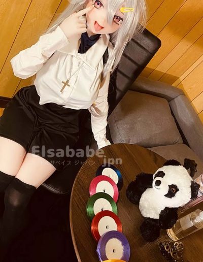 Cosplayer with a silver wig and nun-inspired outfit sits at a table, making a playful face while pointing to their cheek; colorful tapes and a panda plush sit nearby on the table.
