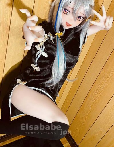 Cosplayer with long silver hair and blue streaks in a black embroidered outfit, crouching against wood panels and reaching toward the camera with a playful expression.